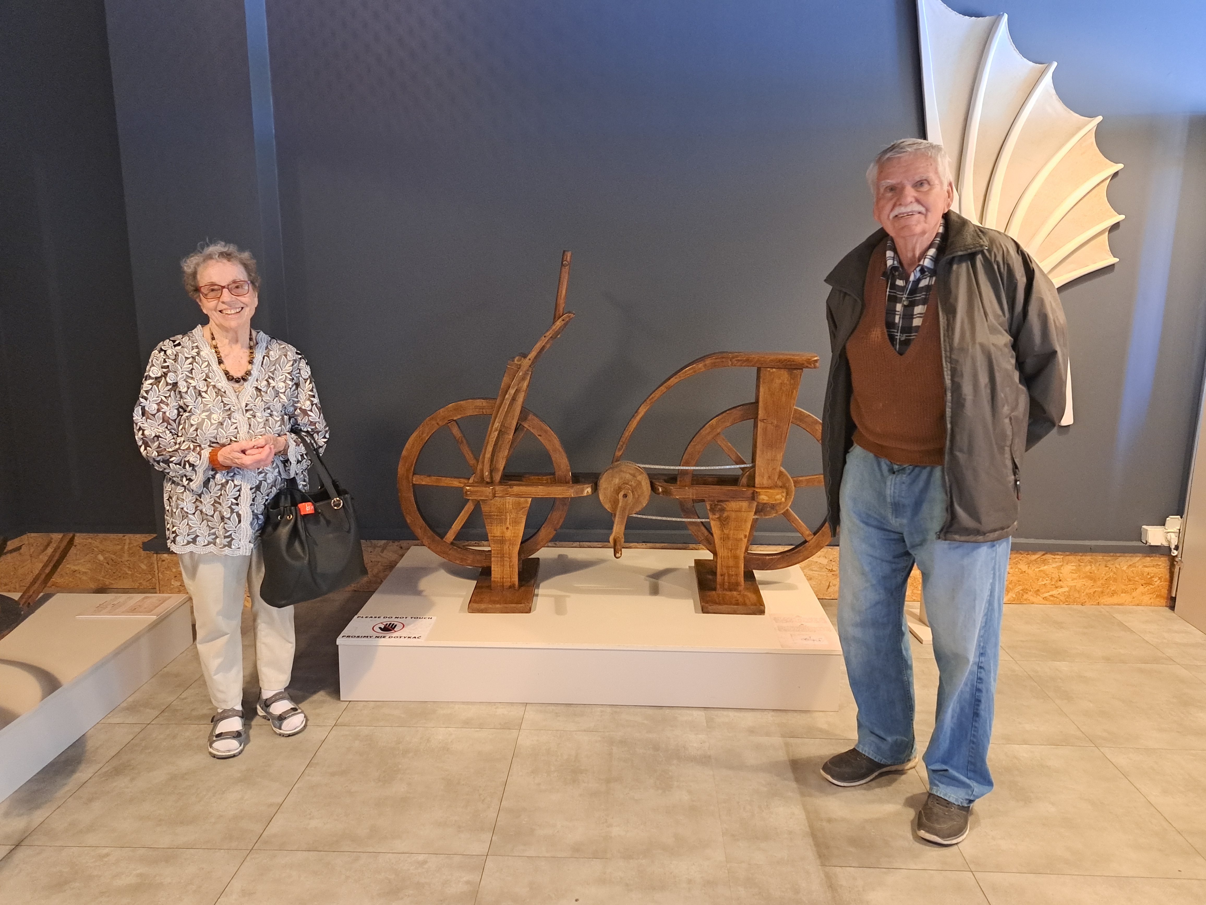 Senior i Seniorka stoją po dwóch stronach maszyny przypominającej rower, wykonanej z drewna
